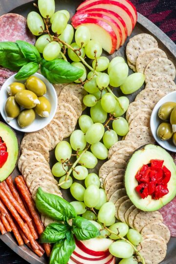 A gluten and dairy-free charcuterie board with fruits, olives, avocado, roasted red peppers, meats, and rice crackers.